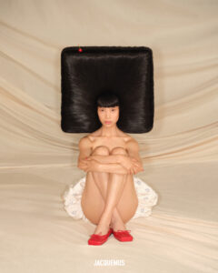 A person sits with knees drawn to the chest on a light, draped fabric background. The individual wears red shoes and is partially covered with a white garment. Notably, the black hair is fashioned into a large, square, flat shape extending outward, creating a striking geometric contrast with the surroundings.