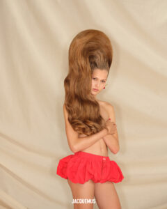 A person stands with an exaggeratedly voluminous hairstyle towering above their head, cascading in large waves. Their expression is calm, and they pose with arms partially crossed over their torso. They wear a bright, short, ruffled skirt. The background is a neutral fabric, draped in soft folds, creating a contrasting backdrop for the vibrant elements of the scene.