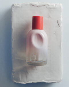 A sleek, translucent bottle with a white, frosted appearance rests elegantly on a textured, rectangular base. The bottle features a distinct, circular indentation, harmoniously blending into its design. Topped with a vibrant red cap, the object exudes minimalistic sophistication, contrasting with the smooth, matte surface beneath. Soft shadows accentuate the bottle's curves, enhancing its refined aesthetic appeal.