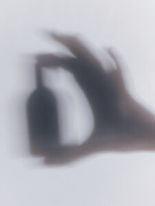 A blurred silhouette showcases a hand gracefully holding a rounded object, likely a bottle, against a soft gradient background. The overall composition suggests an ethereal and mysterious atmosphere, with gentle shadows and indistinct lines adding to the abstract and dreamy quality. The muted tones contribute to the peaceful yet enigmatic feel, inviting contemplation.