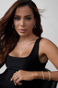 A woman with flowing brown hair and subtle makeup gazes confidently, wearing elegant silver jewelry including hoop earrings, layered necklaces, a bracelet, and rings. She dons a sleek black sleeveless top, exuding style and poise. Her posture is relaxed yet assured, reflecting a modern and sophisticated aesthetic in a simplistic, neutral-toned background.