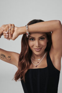 A person with long, wavy hair poses with one arm raised, showcasing their jewelry. They wear multiple layered necklaces and bracelets adorned with charms. The person is dressed in a sleeveless black top. Their expression is confident and relaxed, highlighted by a subtle smile. The background is plain and light, emphasizing the overall composition.