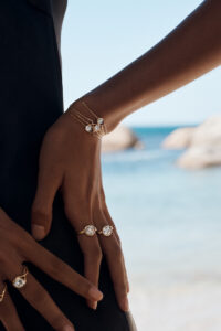 A person stands near a serene beach, one hand gently resting on their hip. They wear elegant gold jewelry featuring shining gemstones on their fingers and wrists. The background reveals a soft-focus view of the sea and rocks under a clear sky, creating a tranquil and luxurious atmosphere. The composition highlights simplicity and elegance.