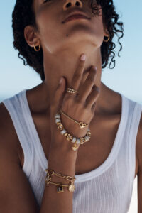 A person with curly hair wears a white sleeveless top, adorned with various gold jewelry. The individual sports hoop earrings and several rings. A charm bracelet and beaded bracelet grace the left wrist, while a delicate necklace wraps around the neck. The pose is introspective, against a clear sky, giving a sense of elegance and simplicity.