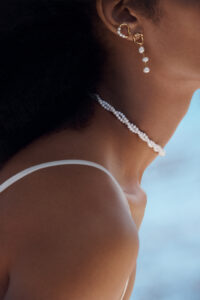 A close-up of a woman's neck and shoulder showcases elegant jewelry. She wears a delicate pearl choker and a pair of drop earrings with gold tones and dangling pearls. Her dark, textured hair partially frames her face, which is turned away from the viewer. Soft lighting highlights her smooth skin and the simplicity of her white strapless top.