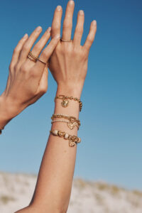Two hands are gracefully raised against a clear blue sky. The wrists are adorned with elegant gold bracelets featuring various designs, including hearts and charms. Simple rings adorn the fingers, adding to the sophisticated look. The background shows a soft-focus landscape with natural, muted colors, enhancing the serene and stylish atmosphere of the scene.