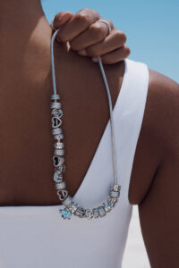 A hand gently holds a charm bracelet, featuring various intricate silver beads and charms, including heart and animal shapes. The bracelet drapes over a shoulder, contrasting with a white sleeveless top. The background is a clear blue sky, enhancing the elegant simplicity of the scene and highlighting the polished details of the jewelry against the skin.
