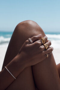 A serene beach scene features a pair of relaxed, sun-kissed legs against a backdrop of gentle waves and a clear blue sky. Adorned fingers and wrist glow with elegant rings and a bracelet, capturing the sunlight. The composition evokes a sense of tranquility and luxury, highlighting the interplay between natural beauty and sophisticated adornment.