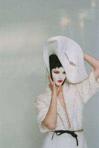 A person wears an extravagant, oversized white hat adorned with an ornate pin, paired with a textured, elegant white outfit. Their face is painted white, highlighted by bold black eyeliner and striking red lipstick. The ensemble is completed with a black and gold belt, creating a dramatic contrast. The background is minimalist, enhancing the avant-garde aesthetic.