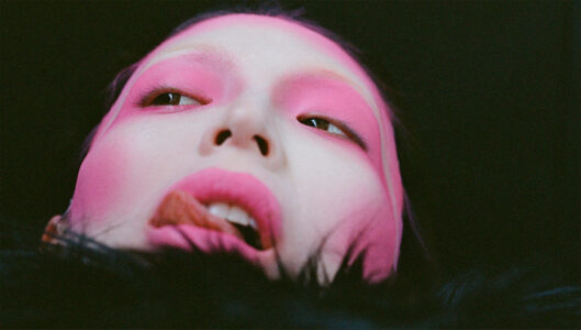 A close-up features a person with vibrant pink makeup covering their eyelids, cheeks, and lips. The upward angle accentuates their serene expression, with their eyes partially closed, and a subtle light shining from above. Soft textures frame the face, adding an ethereal quality against a dark, minimalist background, emphasizing the dramatic makeup and contemplative atmosphere.