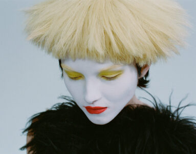 A person with striking, avant-garde style features prominently. Their look is characterized by a voluminous, bleached blond hairstyle and striking makeup. The face is painted stark white, complemented by bold yellow eyeshadow and vibrant red lips. They wear a textured black garment, creating a high-contrast, artistic appearance that exudes a sense of modern, edgy fashion.