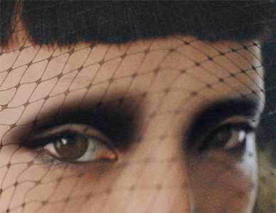 A close-up portrait showcases an individual with a dark, dramatic eye makeup. The gaze is intense, framed by a patterned mesh veil that covers the forehead and partially obscures the face, adding an element of mystery. The eyebrows are well-defined, and the scene is softly lit, emphasizing the texture of the veil against the skin.