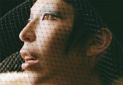 A profile view showcases a person's face partially covered by a delicate black netting. The net casts subtle shadows across the skin, accentuating the eyes and lips. The individual gazes upward, with a calm and contemplative expression. Soft lighting highlights the smooth complexion and the intricate pattern of the net, creating a mysterious, elegant atmosphere.