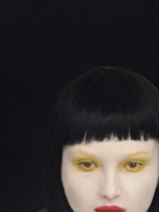 A striking portrait features a person with a sharp black bob haircut and vivid makeup. Bright yellow eyeshadow surrounds the eyes, contrasting with deep red lips, lending an avant-garde and bold look. The pale complexion intensifies the vibrant colors, set against a dark background that enhances the dramatic style and allure of the subject.