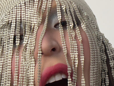 A creative display features a close-up of a face adorned with glossy, silver eye makeup and vibrant pink lips. Overlapping the face are cascading, shimmering strands of sequins, adding an avant-garde feel. These reflective elements create an intriguing contrast with the natural features, combining elements of glamor and modern art in a captivating manner.