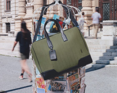A stylish green and black tote bag hangs prominently on a display rack filled with colorful postcards. The background features an ornate building with detailed stonework, large wooden doors, and steps. People pass by, their motion blurred, suggesting a bustling urban environment. The composition highlights the bag's sleek design against the lively street scene.