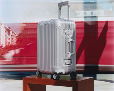 A sleek silver suitcase stands on a wooden platform. It features a ribbed design with secure locks and a retractable handle. Four sturdy wheels add mobility. The background shows abstract blurs of red and hints of urban architecture, suggesting movement and travel. The polished suitcase shines under soft lighting, offering a modern, sophisticated look.