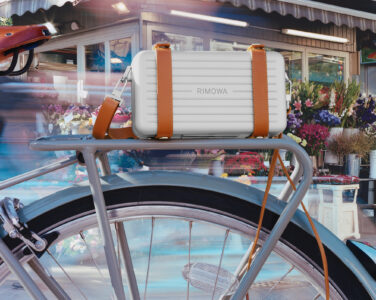 A sleek, modern bicycle is depicted with a white, hard-shell case strapped to the rear carrier using brown leather straps. The blurred background indicates a flower shop, showcasing vibrant blooms. The scene suggests movement and urban lifestyle, capturing a moment of commuting or leisurely riding through a bustling, colorful city environment.