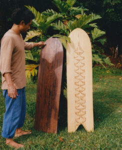 A person stands on lush green grass, examining two elegantly crafted wooden boards. One board is dark with a smooth surface, the other is lighter with intricate carvings. Behind them, vibrant green leaves from a plant create a natural backdrop, emphasizing the craftsmanship and organic setting. The scene exudes a sense of connection to nature and artistry.