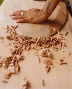 A hand rests on a wooden surface, surrounded by wood shavings. The forearm has a tattoo, and a ring adorns the finger. The scene suggests an ongoing woodworking project, with the result of hand-carving evident in the scattered spirals of shavings. The lighting highlights the wood's natural texture, emphasizing craftsmanship and manual labor.