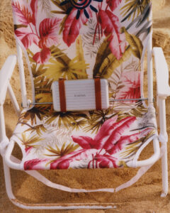 A beach scene showcases a floral-patterned chair nestled in golden sand, capturing a relaxed, summery atmosphere. The design highlights vibrant pink, green, and white tones, reminiscent of tropical settings. A silver, compact suitcase with brown leather straps rests on the chair, suggesting travel or leisure activities, evoking wanderlust and a desire for relaxation and adventure.