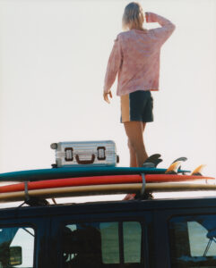 A person stands on the roof of a vehicle, gazing into the distance. They wear a pink long-sleeve shirt and dark shorts. Beside them, a silver suitcase and three surfboards, each in bright colors, are secured to the roof. The setting suggests a beach outing, with clear skies overhead and the reflection of water visible in the car window.