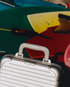 A sleek, silver suitcase with a ribbed design and sturdy handle is placed in the foreground. In the background, a vibrant array of colorful surfboards in shades of green, yellow, red, and blue, with visible fins, create a dynamic and lively composition. Shadows and reflections add depth and contrast to the overall scene.