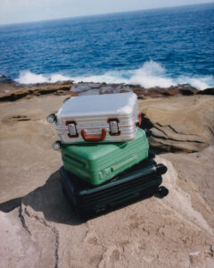 Three suitcases are stacked on a rocky shore, overlooking a vast, vibrant blue ocean. The top suitcase is silver with red handles, the middle one is green, and the bottom is black. Waves crash against the rocks under a clear sky, creating a scenic backdrop of contrasting textures and colors, blending natural beauty with human-made objects.