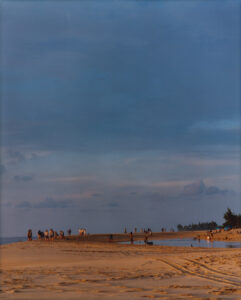 A serene beach scene captures the essence of a calm, early evening. The sky displays shades of blues and soft hues, meeting the horizon where gentle waves kiss the sandy shore. Small groups of people wander along the beach, enjoying the tranquil surroundings. The distant trees create a natural frame, enhancing the picturesque coastal landscape.