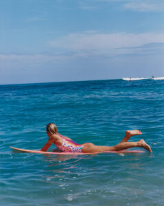 A person lies stomach-down on a surfboard in the ocean, wearing a colorful, floral swimsuit. The calm, blue sea stretches out to the horizon beneath a bright, clear sky. Gentle waves ripple around the surfboard as a distant surfer appears on another wave. The scene captures the essence of a peaceful day at the beach.