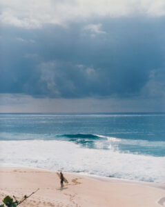 A lone surfer stands on a sandy beach, holding a surfboard, with footprints trailing behind. The ocean waves gently crash on the shore under a vast, cloudy sky. The scene conveys a serene, reflective moment, balancing the turquoise sea's vibrancy with the moody, overcast atmosphere above, hinting at upcoming rain.