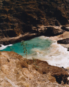 Turquoise waves gently lap against a secluded sandy beach, surrounded by rugged cliffs. People are seen enjoying the water, swimming and wading close to the shore. Two individuals walk along the sandy edge, contrasting against the rocky backdrop. Sparse vegetation clings to the rocky terrain, enhancing the tranquil and remote atmosphere of this coastal paradise.