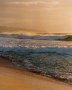 Golden sunlight bathes a vast ocean landscape, highlighting waves as they roll towards a sandy shore. The water reflects hues of orange and yellow, creating a tranquil yet powerful scene. In the distance, mist rises gently, merging with a hazy horizon. The entire setting evokes a serene and picturesque coastal view, inviting peace and contemplation.