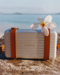 A sleek, rectangular case with horizontal grooves is adorned with brown leather accents. A delicate white plumeria rests on top, adding an elegant touch. The background features a serene beach scene, with soft sand and gentle waves under a clear blue sky, creating a tranquil and luxurious atmosphere.