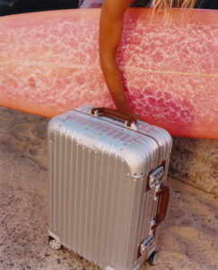 A silver, hard-shell suitcase stands upright on sandy ground, featuring leather accents and a sleek design. Nearby, a person with sun-kissed hair holds a large, smooth pink surfboard with a textured surface. The setting suggests a coastal environment, blending travel and leisure with a stylish and adventurous vibe. The colors and materials create a contrast against the sandy backdrop.