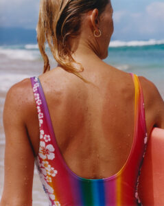 A person stands on a beach, facing the ocean. Their back is visible, with droplets of water glistening on sun-kissed skin. They wear a vibrant, rainbow-hued swimsuit adorned with white floral patterns and hold a pink surfboard. Blond hair drapes down from a ponytail, and blue skies with wispy clouds frame the background, complementing the serene seascape.