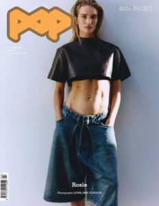 A woman stands against a plain, softly lit wall, her posture relaxed yet self-assured with hands tucked into oversized denim shorts. She wears a structured black cropped top that contrasts with her exposed midriff. Her hair is slicked back, framing a composed, direct gaze. The minimal setting and clean lighting emphasize form, texture, and a quiet, modern confidence.