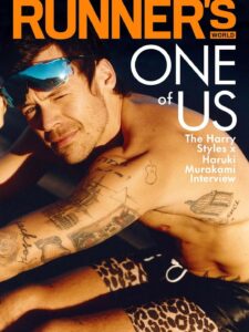 A man with tousled hair lounges shirtless in warm, golden light, his tattooed arms and torso prominently visible. He squints slightly, wearing reflective sport-style sunglasses pushed up on his face. Leaning forward with a relaxed, playful expression, he wears patterned shorts, while the close framing and sunlight create an intimate, sun-soaked, athletic mood.
