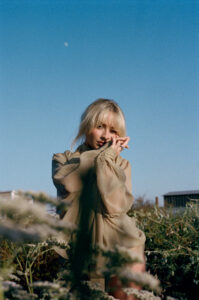 A woman stands in a sunlit field, surrounded by tall wild plants. She wears a sheer, long-sleeved, beige dress that catches the light. Her blonde hair is loose with bangs framing her face. With one hand raised near her eyes, she gazes directly ahead. The sky above is clear blue with a faint moon visible overhead.
