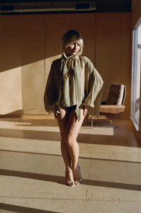 A person stands indoors, bathed in warm, soft light. They wear a sheer, loose-fitting top and dark shorts. Their hair appears tousled, casting soft shadows on their face. A window on the right allows natural light to fill the room, while a modern chair sits in the background. The setting exudes a relaxed, tranquil atmosphere.