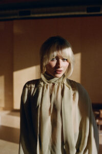 A woman with light blonde hair and wispy bangs stands in warm, directional sunlight that casts strong shadows on the wall behind her. She wears a flowing, semi-sheer beige blouse with soft pleats and a high neckline tied loosely at the throat. Her expression is slightly parted, caught between curiosity and contemplation, creating a quiet, cinematic moment within a minimal interior.