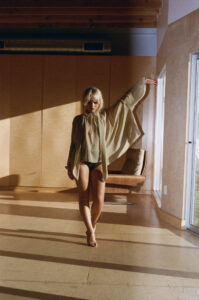 A person stands gracefully on a sunlit floor, adorned in a sheer, flowing garment with extended sleeves. The space features wooden elements and soft shadows, with a minimalist interior and a large window allowing natural light to create a serene atmosphere. The overall ambiance feels calm and contemplative, highlighting the blend of natural and architectural elements.
