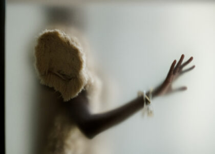 A blurred figure is captured with an arm extended, fingertips splayed against a frosted glass surface. The person wears a fluffy, cream-colored garment. A bracelet with small charms is visible on the wrist, creating a soft yet striking visual contrast against the pale, muted background, which enhances the mysterious, ethereal quality of the scene.