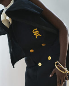 A stylish ensemble showcases a tailored dark jacket adorned with gold buttons and a striking yellow emblem. The sophisticated look features a structured, layered shoulder detail and an elegant white scarf or tie. Completing the ensemble is a bold, overlapping gold bracelet on the wrist, adding a touch of luxury and refinement to the sophisticated attire.