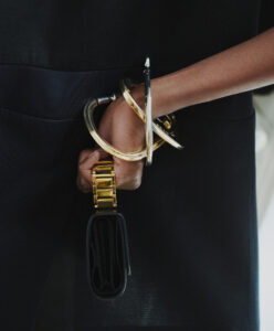 A person holds a small, black rectangular bag with a gold chain. The hand is adorned with several thick, shiny gold and silver bangles. The background features a dark fabric, possibly a garment, adding contrast to the metallic accessories. The composition highlights the interplay between the elegant jewelry and the minimalistic accessory.