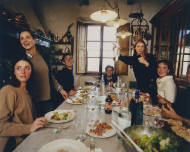 A group of people gather around a dining table enjoying a meal together. Plates of food and bottles are spread out on the table, and the setting features warm, inviting lighting. The room has rustic decor, with wooden shelves and a window letting in natural light. One person gestures with a bottle, creating an energetic atmosphere.
