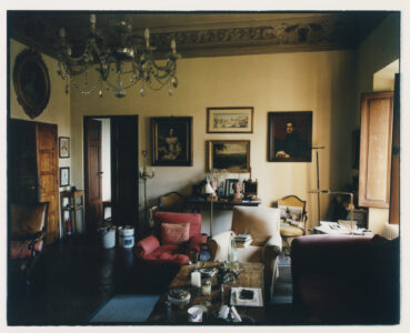 A vintage living room features classic decor with elegant chandeliers and ornate ceiling details. Rich wooden furniture and plush armchairs create a cozy ambiance. The walls are adorned with framed paintings and portraits, adding an artistic touch. Sunlight gently streams through a window, illuminating a coffee table cluttered with books and knickknacks, enhancing the room's warm and inviting atmosphere.