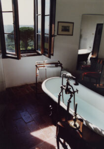 A serene bathroom features a vintage-style bathtub with elegant fixtures on a tile floor. Natural light streams through open wooden windows, highlighting the picturesque view of green trees and distant hills. A wooden chair and towel rack complement the minimalist decor, while a large mirror reflects the peaceful, airy atmosphere, creating a sense of spaciousness.