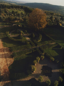 A beautifully manicured garden features intricate hedge patterns and pathways, bordered by terracotta pots of vibrant plants. The lush greenery leads to a small ornamental pond at the center. In the background, rolling hills and dense woodland stretch into the distance. A large tree with autumnal leaves adds a splash of color to the serene landscape.
