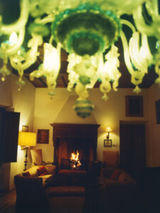 A cozy living room features a glowing, ornate chandelier in the foreground. Comfortable sofas and armchairs encircle a wooden coffee table. In the background, a warm fire crackles in a grand fireplace, flanked by elegant wall lamps and artwork. The setting exudes warmth and comfort, ideal for relaxation and intimate gatherings in an inviting ambiance.